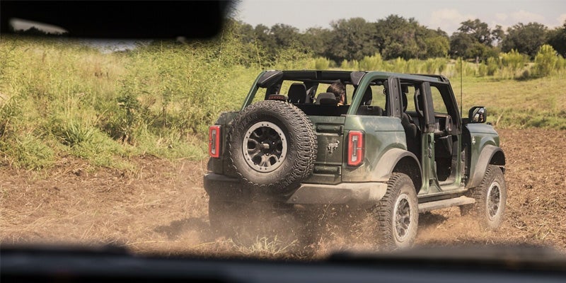 Rear view of a green 2024 Ford Bronco driving off-road through a dirt path, with spare tire mounted on the back